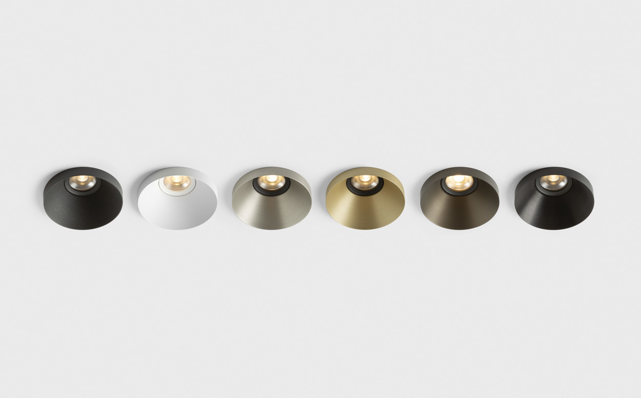 A lineup of six Kup recessed spotlights mounted in a white ceiling, showcasing different finishes. From dark bronze and white to brushed champagne, gold and darker metallic tones, each luminaire features Kup’s distinctive raised-edge design and softly illuminated recessed light source. The image highlights the collection’s material and finish options within a clean architectural setting.