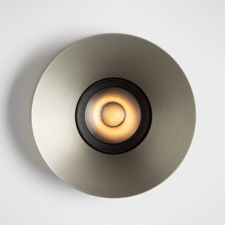 Close-up of Kup, a round recessed spotlight with a brushed metallic finish and distinctive raised edge. The deeply recessed light source emits a warm, precisely controlled beam, highlighting the luminaire's refined cup-shaped design and architectural presence.