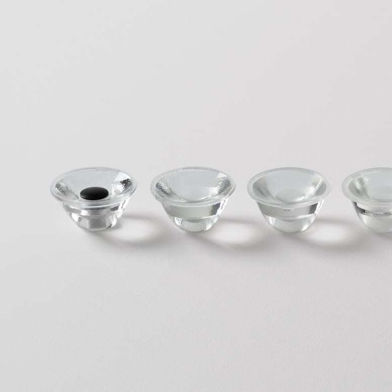 Close-up of four transparent TIR lenses arranged in a row on a white surface. Each lens features a slightly different optical geometry, illustrating the range of beam distributions available within the optical platform. Soft shadows and reflections emphasise the precision-engineered glass components.