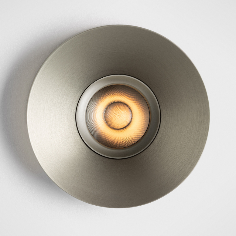 Close-up of Kup, a round recessed spotlight with a brushed metallic finish and distinctive raised edge. The deeply recessed light source emits a warm, precisely controlled beam, highlighting the luminaire's refined cup-shaped design and architectural presence.