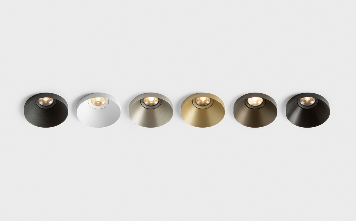 A lineup of six Kup recessed spotlights mounted in a white ceiling, showcasing different finishes. From dark bronze and white to brushed champagne, gold and darker metallic tones, each luminaire features Kup’s distinctive raised-edge design and softly illuminated recessed light source. The image highlights the collection’s material and finish options within a clean architectural setting.